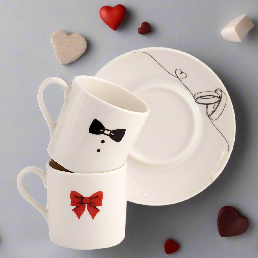 A set of white porcelain coffee cups with black and red decorative elements, accompanied by two saucers with a line drawing of a bride and groom. The cups and saucers are displayed with heart-shaped decorations scattered around.
