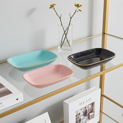 Three rectangular ceramic trays in turquoise, pink, and black on a shelf with a vase and books.