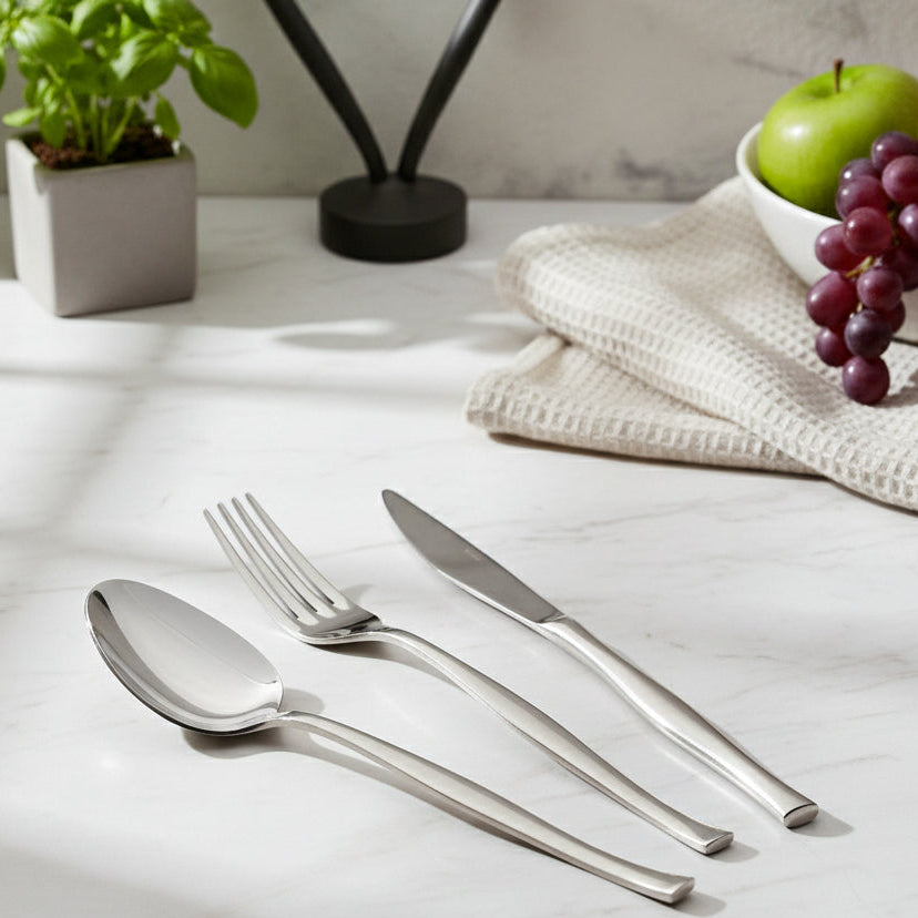 Silver cutlery set on a light wooden surface with fruits and a plant in the background.
