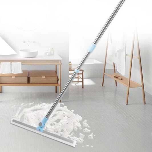 An extendable squeegee with a stainless steel arm and a silicone blade is shown with a white handle and blue accents. It is being used to clean a bathroom floor, leaving a trail of soap behind as it moves across the floor.